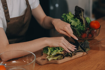 Delicious fruit and vegetables on a table and woman cooking. Housewife is cutting green cucumbers on a wooden board for making fresh salad in the kitchen.