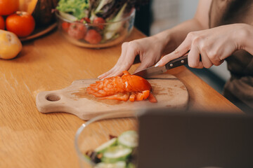 Delicious fruit and vegetables on a table and woman cooking. Housewife is cutting green cucumbers on a wooden board for making fresh salad in the kitchen.