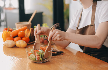 Delicious fruit and vegetables on a table and woman cooking. Housewife is cutting green cucumbers on a wooden board for making fresh salad in the kitchen.