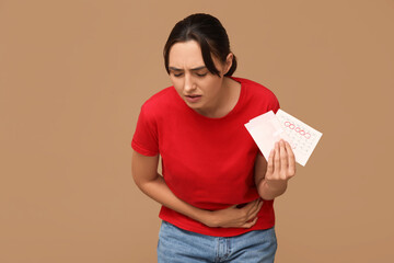 Young woman holding pad and calendar with marked days of menstruation on brown background