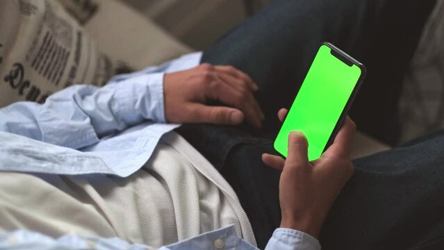 Man holding smart phone with green chroma key screen while sitting on sofa in living room, using Internet, social media, online shopping, watches video or movie