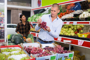 European man choosing grape in greengrocer. Asian woman with cart full of vegetables walking behind him.