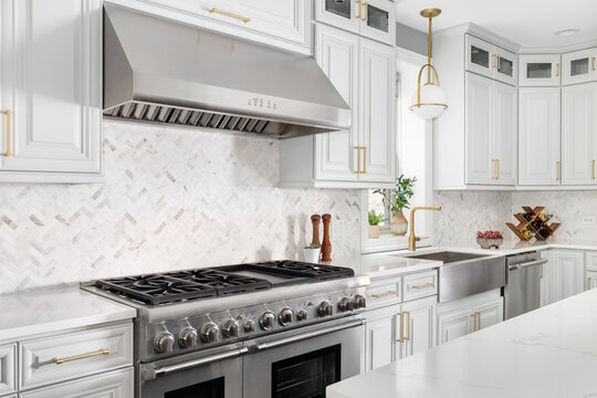 A kitchen detail with grey cabinets, a herringbone tile backsplash, stainless steel apron sink, and a gold hardware and faucet. No brands or logos.