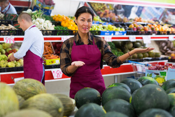 Asian woman in apron standing in salesroom of greengrocer and presenting assortment of fruits and...