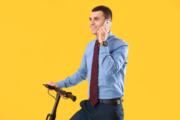 Fototapeta na wymiar Young businessman with electric scooter talking by mobile phone on yellow background