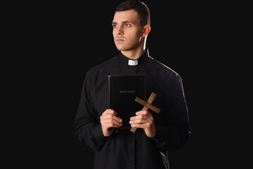 Young priest with Holy Bible and cross on dark background