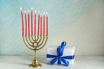 Menorah with burning candles and gift box for Hanukkah celebration on light color background