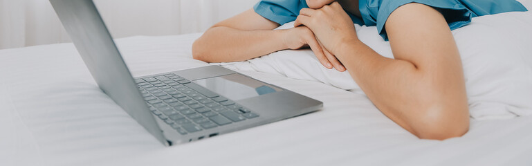 Young Asian woman with beautiful face, long hair, blue shirt lying on bed in white bedroom at home with cup of coffee and video call with laptop talking to relatives on vacation. holiday concept.