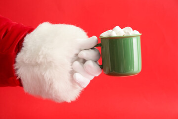 Obraz na płótnie Canvas Santa Claus holding cup of tasty cocoa drink with marshmallows on red background, closeup