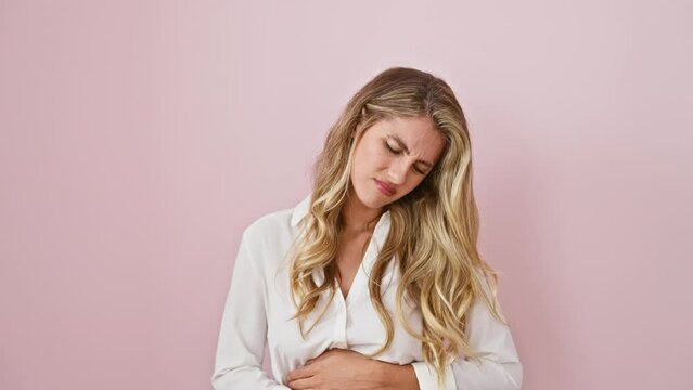 Young blonde woman in pain, hand clutching her stomach over a pink isolated background. wearing shirt, she stands ill, bearing the ache of a severe stomach illness painfully.