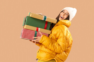 Young woman in winter clothes with Christmas gifts on beige background