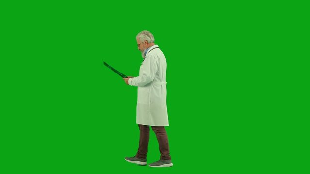 Portrait of aged man medic on chroma key green screen. Side view senior doctor walking through clinic or hospital looking at x-ray picture.