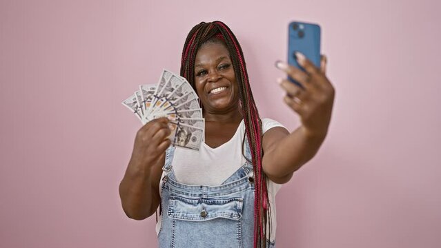 Black woman with braids, beautiful and all smiles, holds up dollars, showing off her win in the business world. she's captured the moment with a selfie over a pink, isolated background.