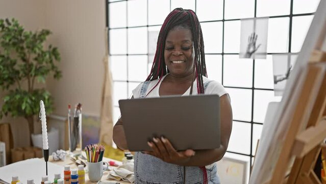 Confident african american woman artist with braids, smiling as she paints on canvas and uses laptop in a cozy art studio
