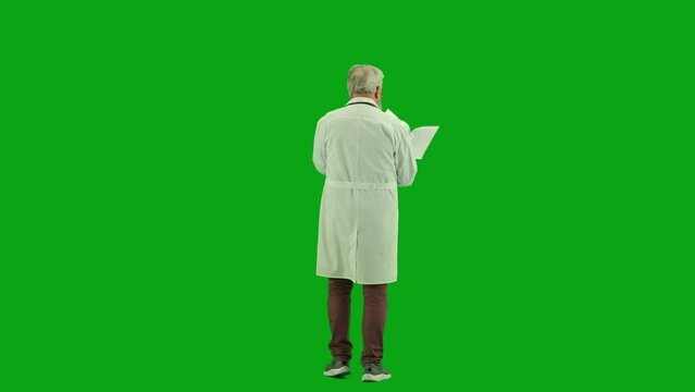 Portrait of aged man medic on chroma key green screen. Back view senior doctor in uniform walking in hospital holding paper documents reading it.
