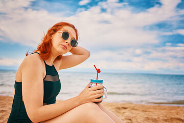 Beautiful woman in swimsuit is sitting on beach with cocktail and look away. Tropical alcohol beverage. Red-haired lady with sunglasses on vacation. Summer day with cloudy sky. Holidays at sea.