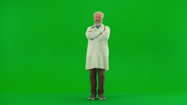 Portrait of aged man medic on chroma key green screen. Senior doctor in white coat standing with crossed arms and smiling at the camera.