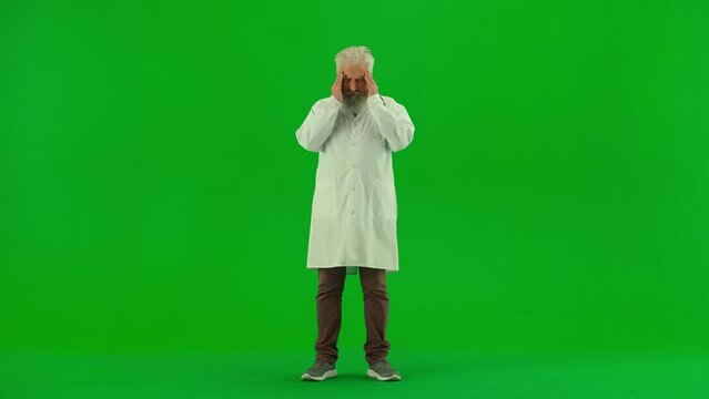 Portrait of aged man medic on chroma key green screen. Senior doctor in white coat standing massaging temples, having headache looking tired.