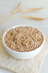 Wheat groats in a white bowl with three ears of wheat on a light background. Rustic style. Concept of healthy food. Vertical orientation.