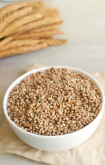 Wheat groats in a white bowl with ears of wheat on a light background. Rustic style. Concept of healthy food. Vertical orientation.