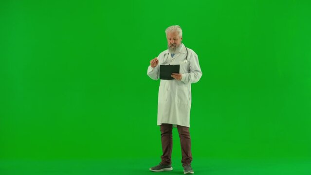 Portrait of aged man medic on chroma key green screen. Senior doctor in white coat with clipboard standing pointing at empty area talking.