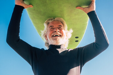 happy senior with surftable on his head is smiling and laughing - old and mature man having fun surfing with a black wetsuits - active retired adult doing activity alone