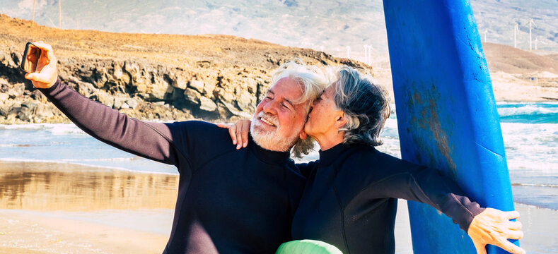 couple of two cute seniors and pensioners doing exercise and taking a selfie with their surftables and wetsuits - woman kissing her husband