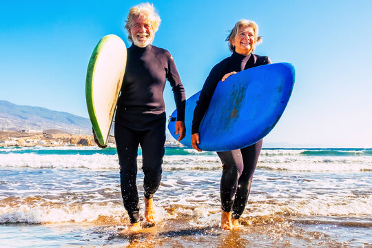 two old and mature people having fun and enjoying their vacations outdoors at the beach wearing wetsuits and holding a surfboard to go surfing in the water with waves - active senior smiling water