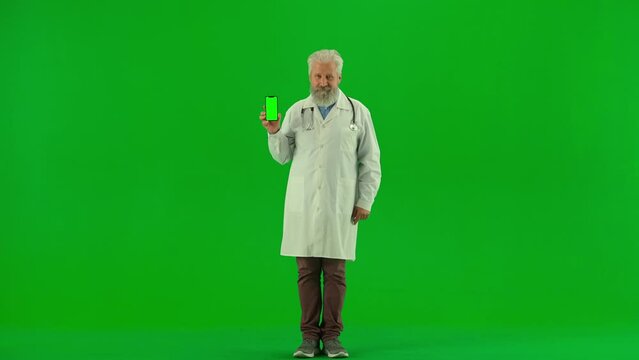 Portrait of aged man medic on chroma key green screen. Senior doctor in coat holding smartphone with workspace mockup, smiling at the camera.