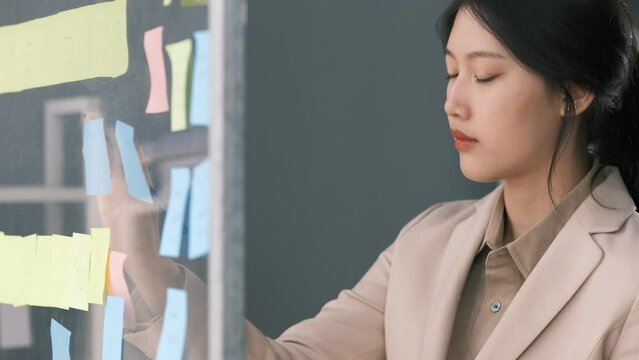 Young smiley attractive businesswoman using sticky notes in glass wall to writing strategy business plan to development grow to success