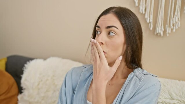 At home, young beautiful hispanic woman in casual clothes, covering her mouth in shock. surprised, afraid for a secret she let slip. expression of fear and silence in her living room.