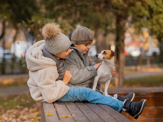 Brother and sister sit in an embrace with a dog on a bench for a walk in the autumn park. Boy, girl and jack russell terrier. 