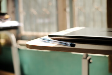 A photo of a cream colored wooden table on which there is a laptop and a blue ballpoint pen,...