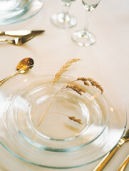 Modern minimalist place setting with clear glass plate, gold flatware, and rustic dried plants