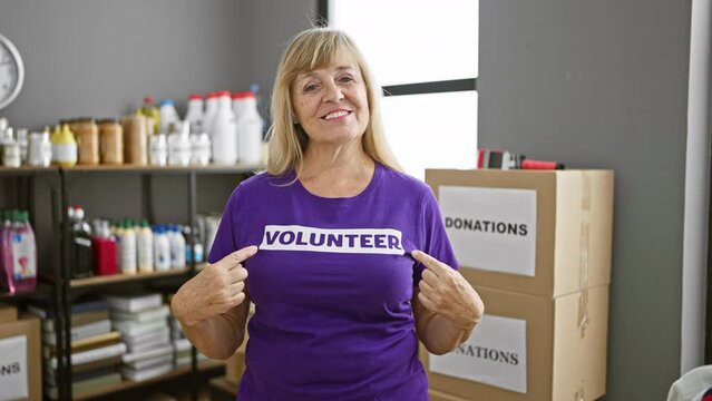 Blonde middle-age woman, full of joy, points at her volunteer uniform while radiating a confident smile at the bustling charity center, feeling unity and altruism in her invaluable service.