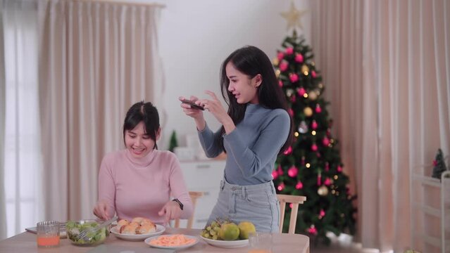 Asian female friends capture memories by taking photos together using smartphones at the dining table in the festive atmosphere of Christmas at her home