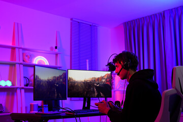Caucasian gamer boy focus on playing fps shooting game as a sniper with his team while using joystick and two monitor screen with wireless headphone gadget hobby lifestyle in purple neon light room © MergeIdea