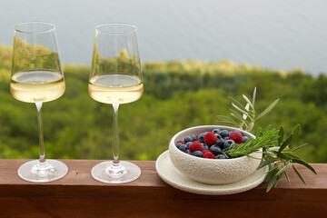 White wine in glasses against a background of blue sky and sea and a plate with raspberries and blueberries next to it . High angle. Copy Space.