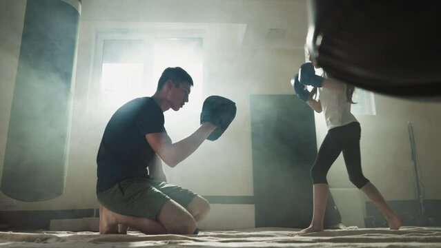 Kid kicks pad with trainer support in combat gym. Kickboxing trainer teaches barefoot little girl leg attack in sports school slow motion. Self-defense lesson