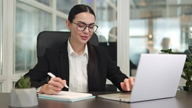 Concentrated office worker sitting at table and working with wireless laptop while taking notes in office. Caucasian calm woman wearing glasses and writing information to notebook in bright room.