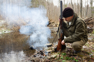 A hunter sits by a dying campfire on the bank of a forest river