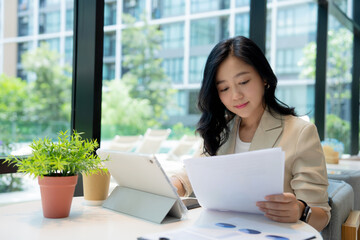 Beautiful Asian businesswoman analyzing charts using calculator and laptop with documents and holding a pen.