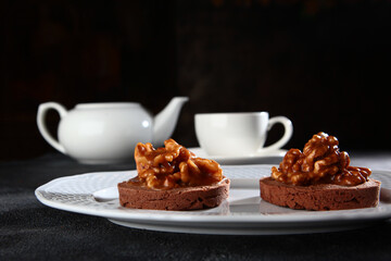 Chocolate brownie with walnuts. The concept of breakfast. The white porcelain teapot is out of focus. Black background.