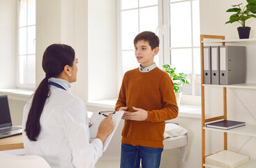 Fototapeta premium Female friendly doctor sitting back talking with a child patient during medical examination and listening complaints in clinic. Pediatrician checking a young boy and giving consultation in exam room.