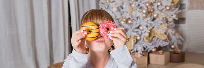 cheerful young woman holds donuts in her hands on christmas tree background, winter holidays. portrait of funny girl in pajamas covering her eyes with donuts