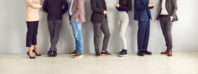 Legs of different business people standing in row and communicating with each other. Unrecognizable people in stylish business and smart casual clothes stand on wall background. Banner. Cropped image. - obrazy, fototapety, plakaty