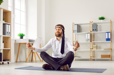 Funny business man or office worker in white shirt, with necktie around his head sitting with crossed legs on yoga mat on floor, doing meditation, relaxing and calming down on stressful day at work