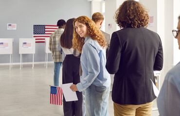 Young happy american female voter standing in a queue at vote center and looking cheerful at camera registering at polling station on USA election day. Woman holding ballot paper at electoral center. - obrazy, fototapety, plakaty