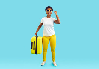 Full length portrait laughing happy african american woman in yellow pants with yellow suitcase in hand showing win gesture on turquoise background. Travelling, tourism, journey, holiday trip concept