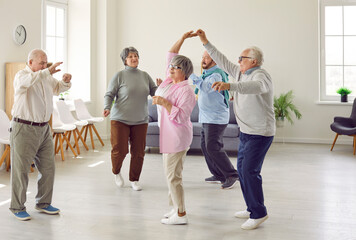 Retirement community. Elderly men and women actively spend time having fun and dancing at party in nursing home. Group of senior Caucasian people laughing and dancing together in living room. - obrazy, fototapety, plakaty